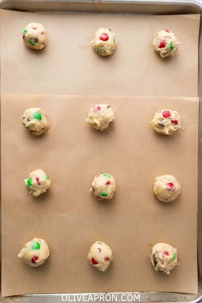 Unbaked Christmas M&M cookie dough balls on a parchment-lined baking sheet, ready to go in the oven.
