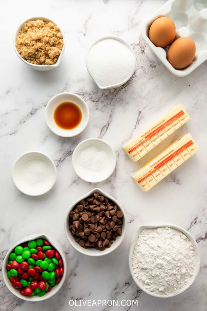 M&M cookie ingredients: butter sticks, eggs, flour, brown sugar, white sugar, vanilla, baking soda, salt, chocolate chips, and red and green M&M’s.
