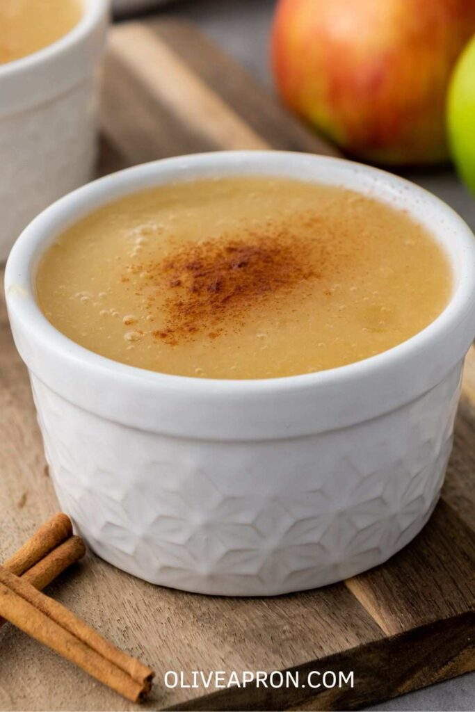 Small white bowl filled with homemade applesauce topped with ground cinnamon, with apples in the background.