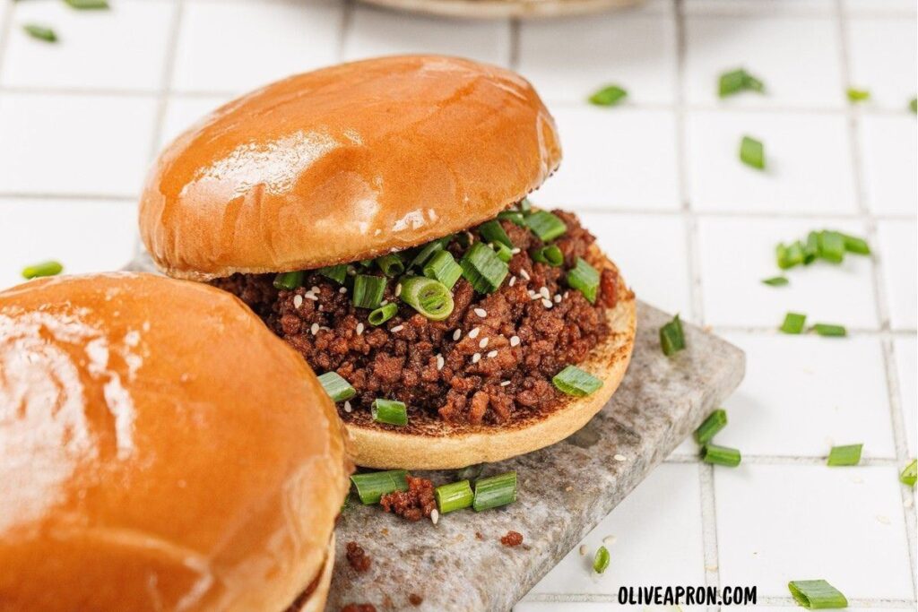 Sloppy Joe sandwich with glossy bun, filled with seasoned beef and scallions, on a cutting board.