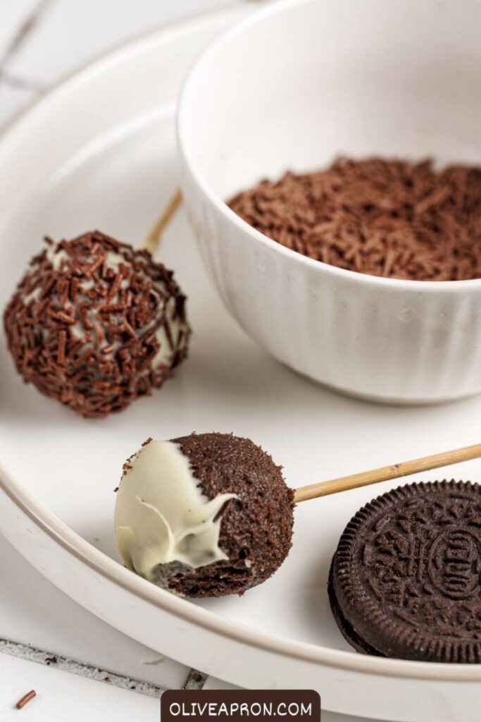 Homemade Oreo cake pops coated in white chocolate and chocolate sprinkles next to a bowl of sprinkles and Oreo cookies.