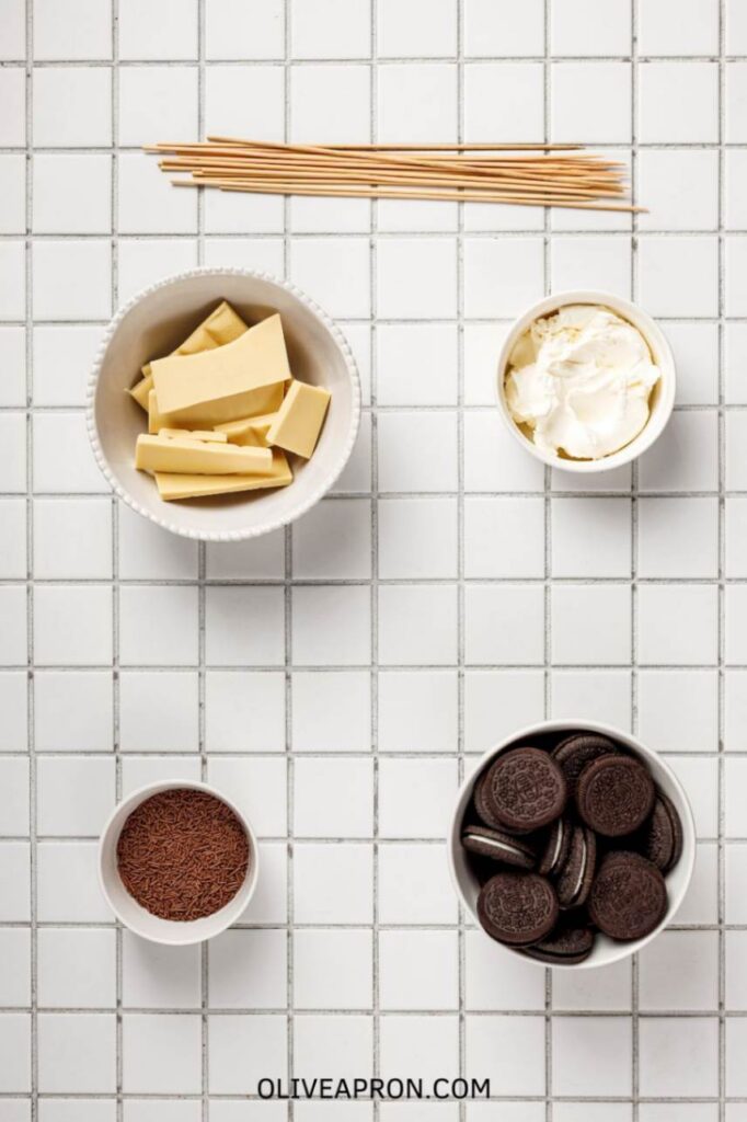 Oreo cake pop ingredients displayed on a tiled surface including Oreo cookies, cream cheese, white chocolate pieces, chocolate sprinkles, and wooden sticks.