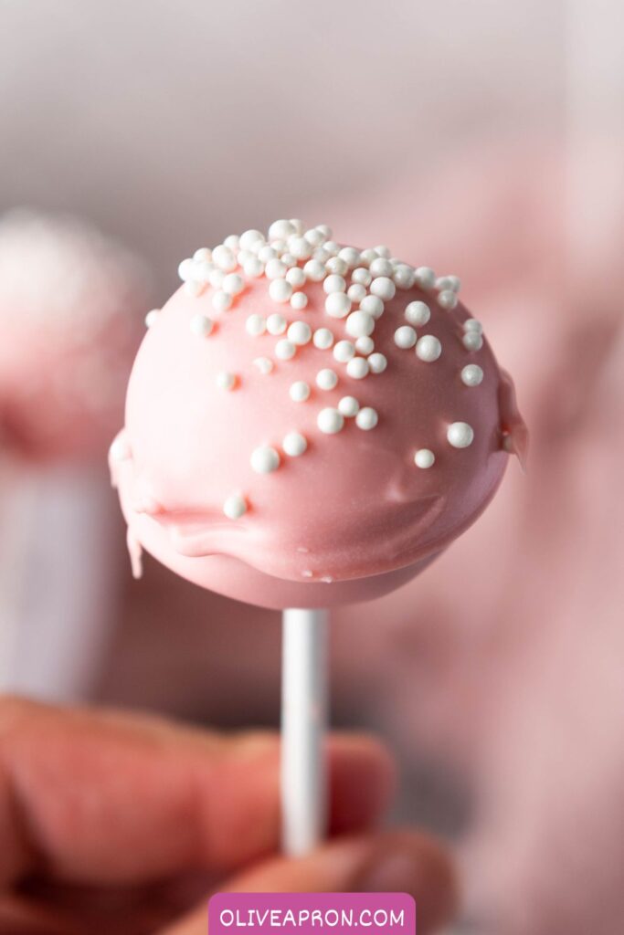 Pink cake pops coated in pastel pink candy melts and topped with white sprinkles, displayed on sticks for a cute and easy cake pop dessert.