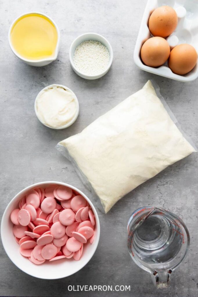 Cake pop ingredients on a grey surface including pink candy melts, boxed cake mix, eggs, oil, water, frosting, and white sprinkles