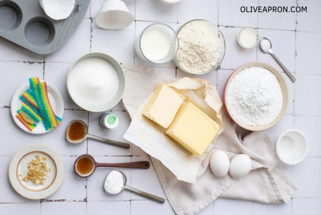 Ingredients for St Patricks Day cupcakes including flour, sugar, butter, eggs, powdered sugar, milk, vanilla, food coloring, and rainbow candy strips arranged on a kitchen surface.