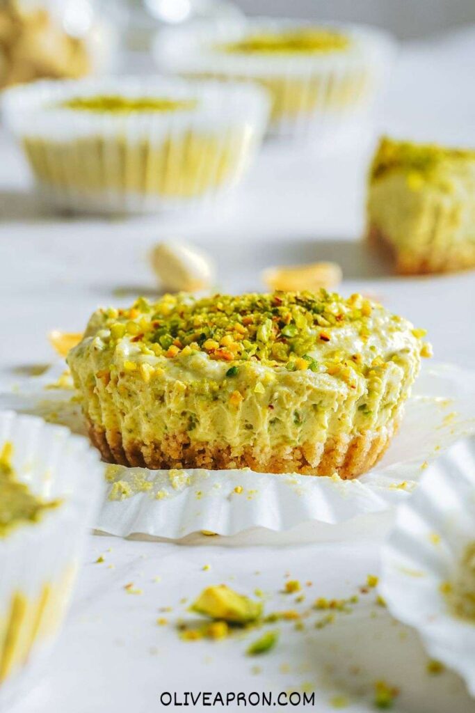 Mini pistachio Greek yogurt cheesecakes over a white surface with scattered pistachios .