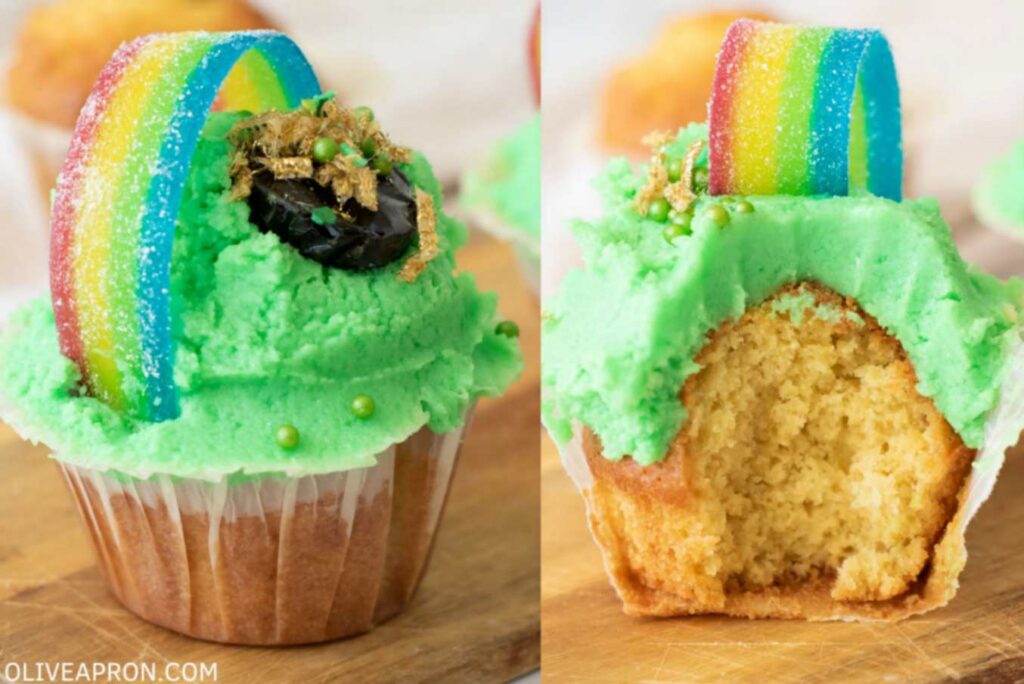 Close up of St Patricks Day cupcakes topped with bright green frosting, rainbow candy arches, and gold sprinkles, including one cupcake with a bite showing the soft vanilla crumb inside.