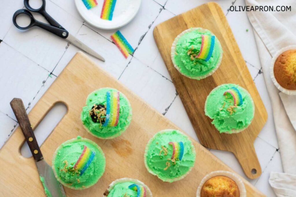 Overhead view of St Patricks Day cupcakes decorated with bright green frosting and rainbow candy strips, arranged on wooden boards with sprinkles and baking tools nearby.