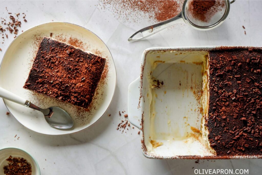 Slice of homemade tiramisu cake served on a plate beside the baking dish, topped with cocoa powder and chocolate shavings.