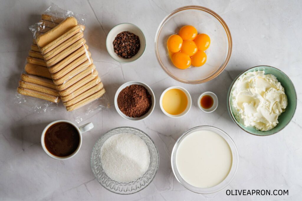 Ingredients for homemade tiramisu are arranged on a countertop, including ladyfingers, mascarpone, espresso, egg yolks, cream, cocoa powder, and vanilla.